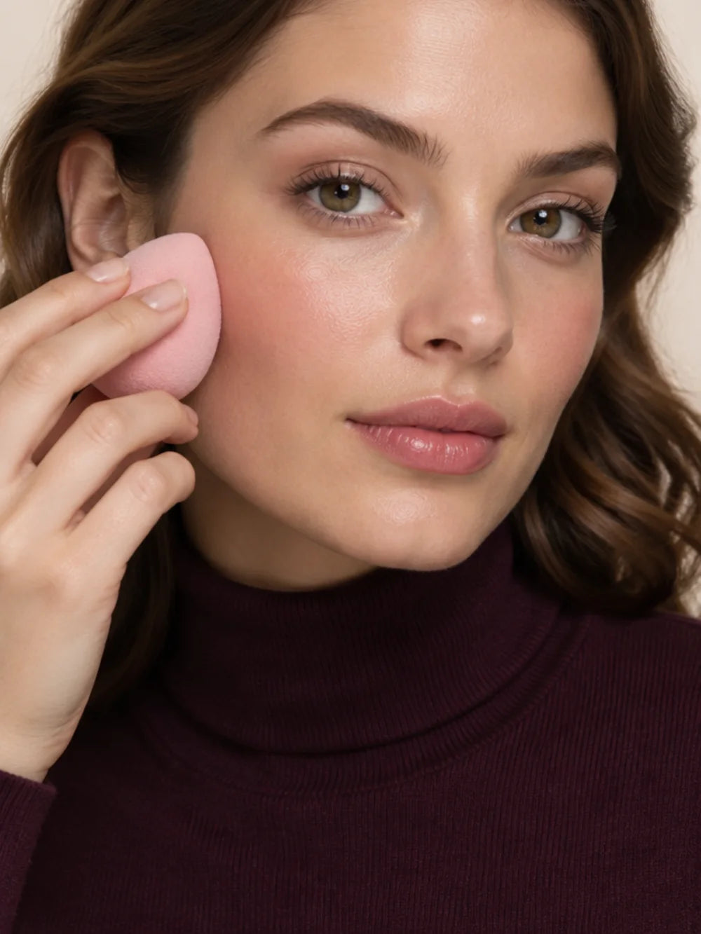 Young woman in a plum turtleneck blending foundation with a makeup sponge